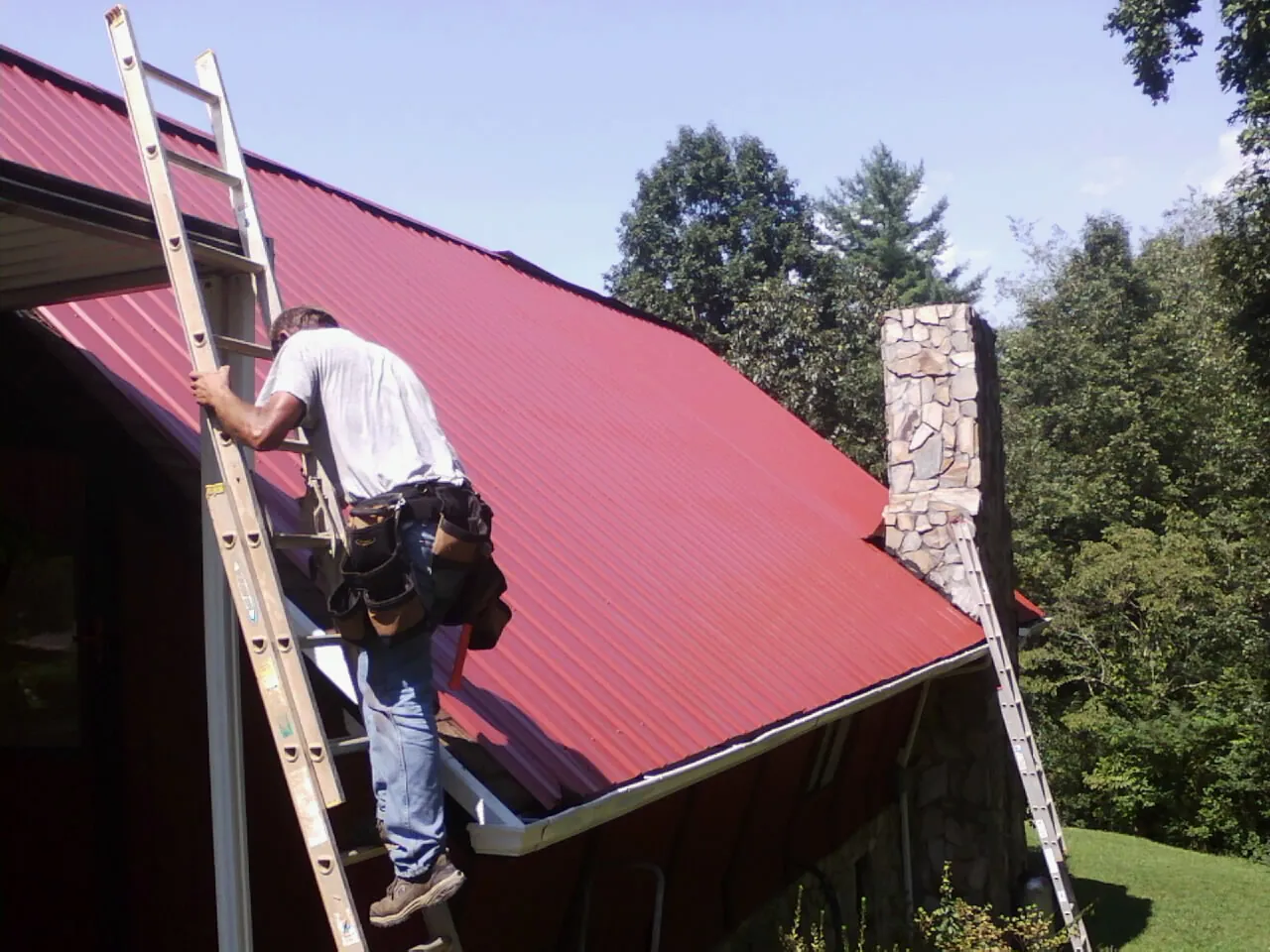Roofer on a ladder beside a red metal roof and stone chimney — Soffit & Fascia Repair by Roofing Brothers in Morven